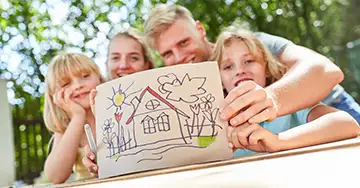 Wissenswertes zum Hausbau Glückliche Familie mit zwei Kindern präsentiert stolz ein selbstgemaltes Bild ihres Traumhauses im Garten.
