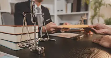 Infos zu Rechtsschutzfragen Anwalt übergibt rechtliche Unterlagen am Schreibtisch mit Justitia-Waage.