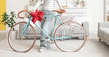 Alles über die private Haftpflicht Eine Person steht hinter einem hellblauen Vintage-Fahrrad, das mit einer großen roten Geschenkschleife am Lenker verziert ist, in einem hellen Wohnzimmer.